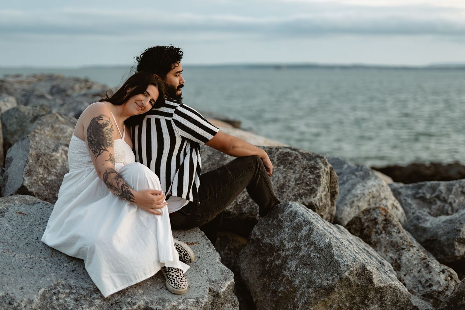 A romantic couple sits on coastal rocks overlooking a serene ocean view at sunset.