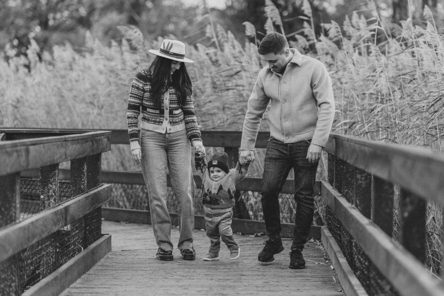Family walking together on wooden boardwalk through tall grass in black and white.