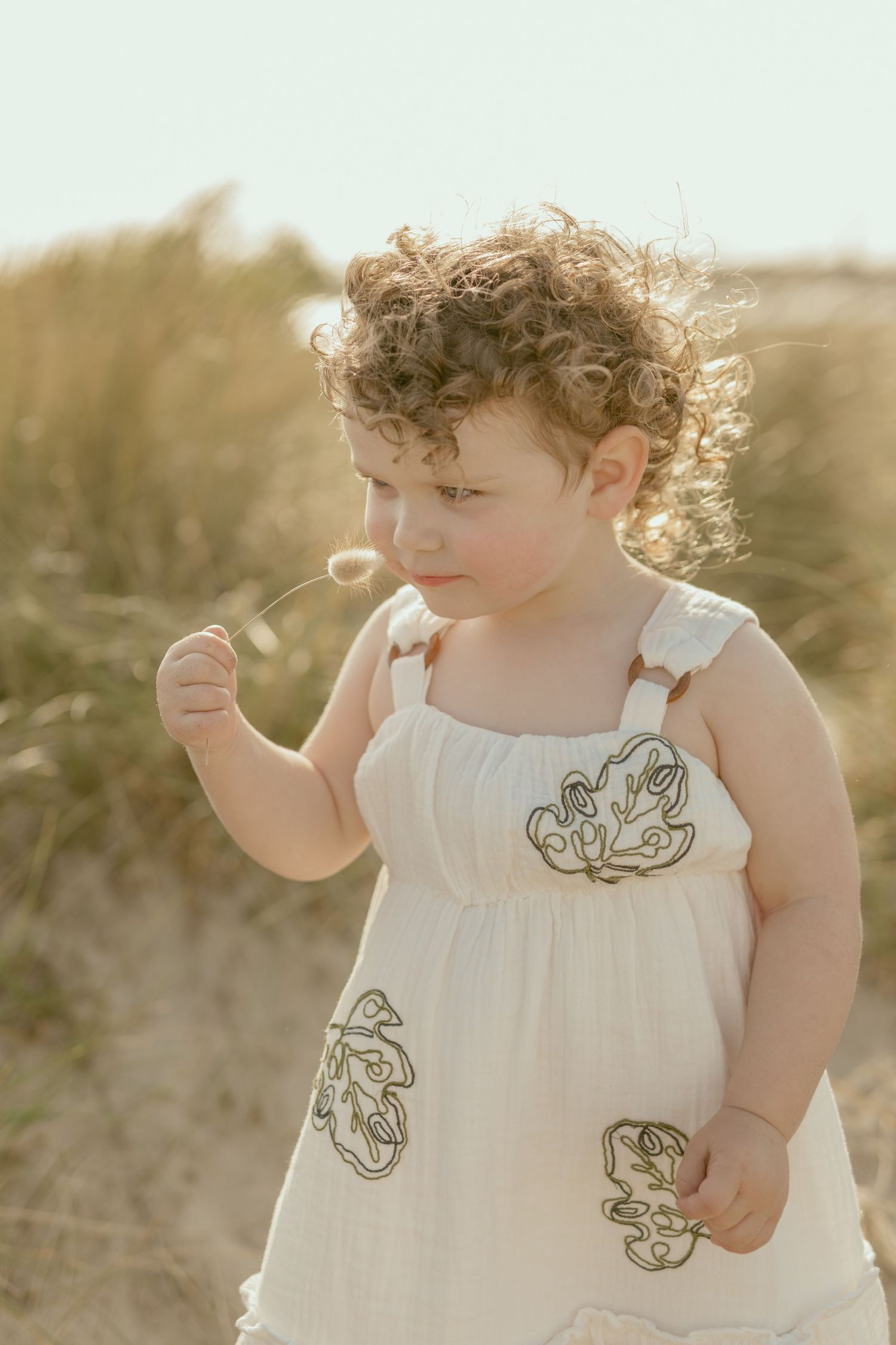 A toddler in an embroidered white dress explores the beach at golden hour.
