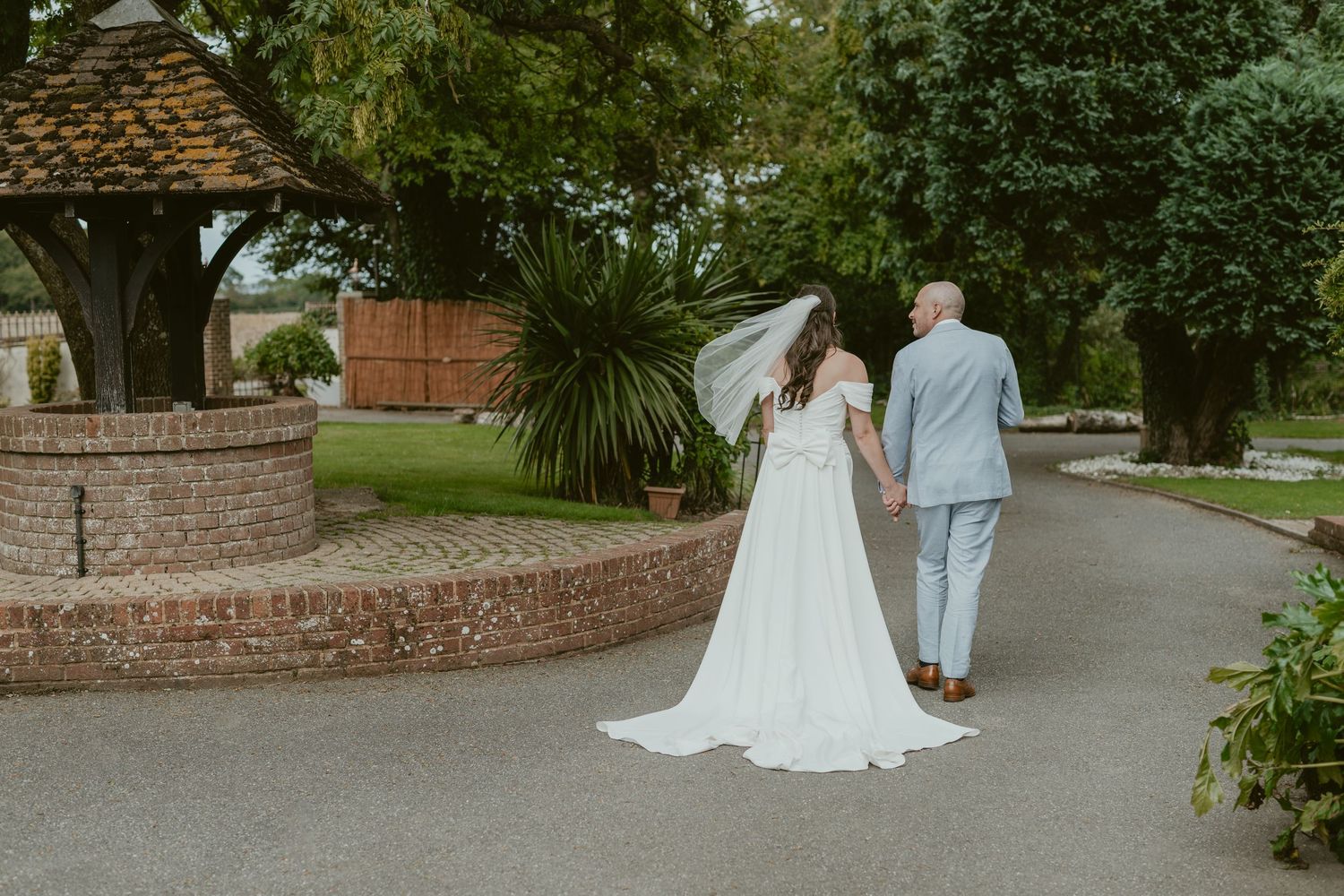 A couple in wedding attire walks along a garden path surrounded by lush greenery.