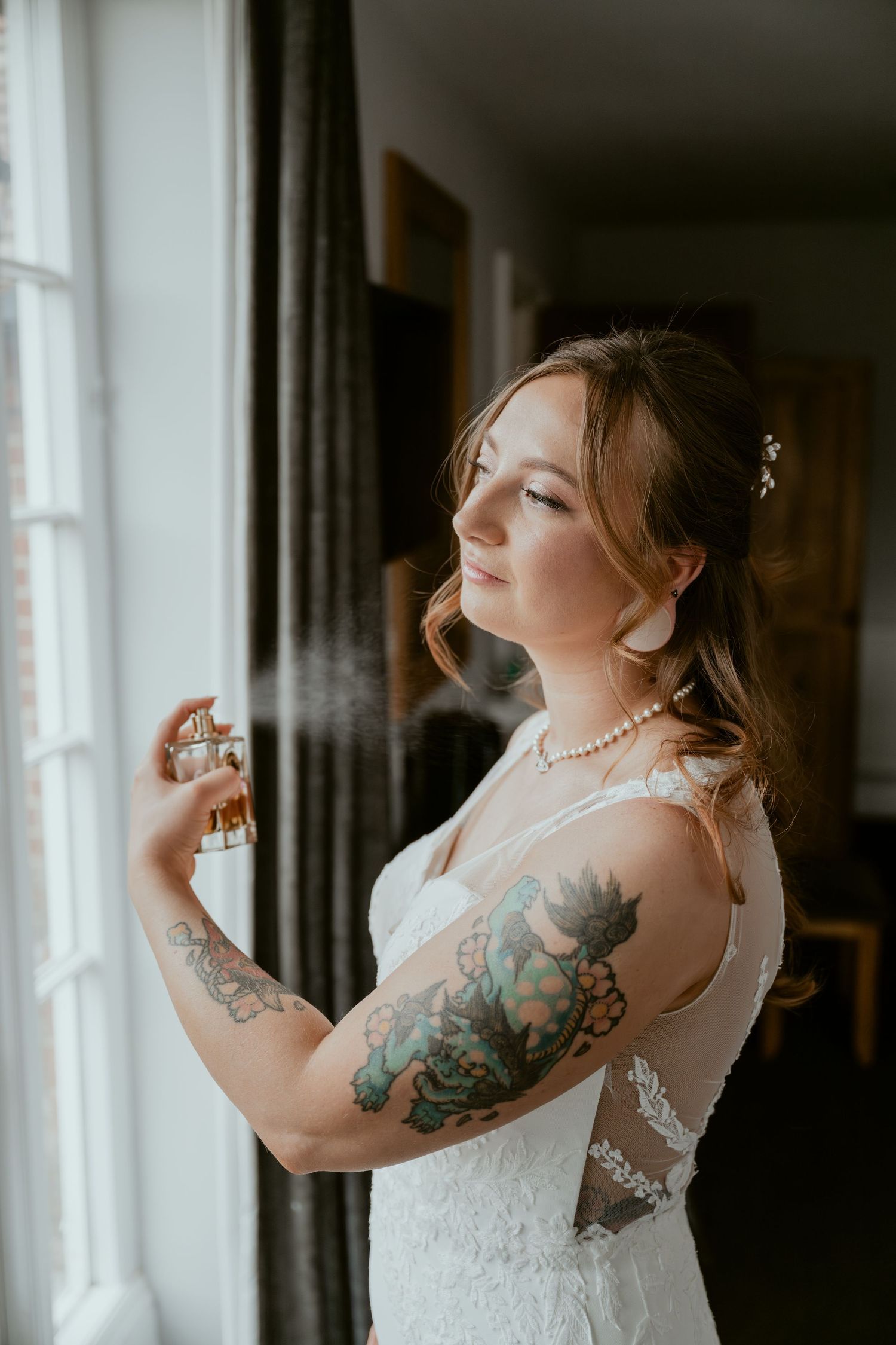 Bride in white lace dress with floral arm tattoos poses gracefully by window in romantic boudoir photoshoot.