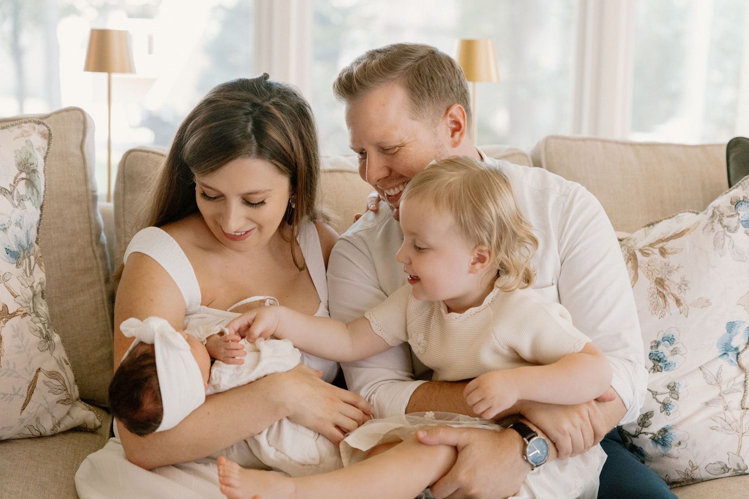 Family cuddles together on cozy white bedding in a sun-lit bedroom.