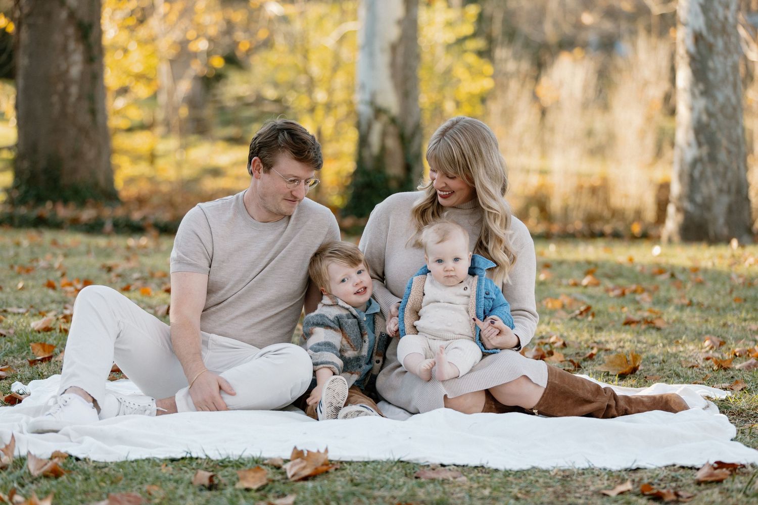 Family lounging together on white blanket during autumn photoshoot with fallen leaves.