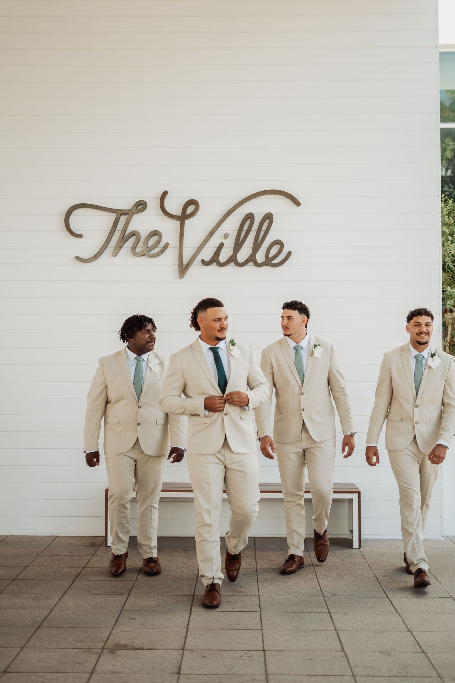 Groomsmen in beige suits pose casually against white wall with rustic signage.