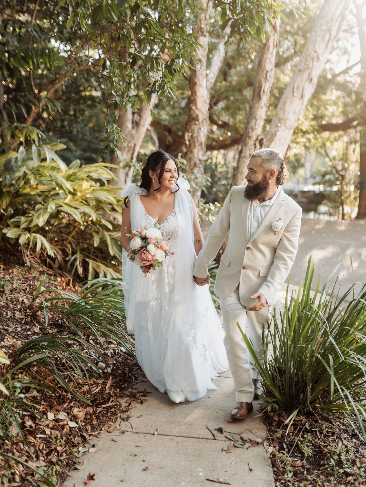 Bride and groom walking hand-in-hand on garden path, she in white gown with bouquet, he in beige suit.
