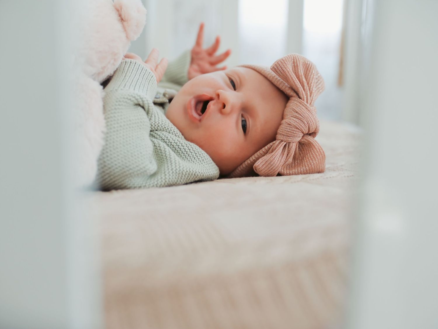 Smiling baby in green onesie and pink bow headband lying in a white crib, reaching up playfully.