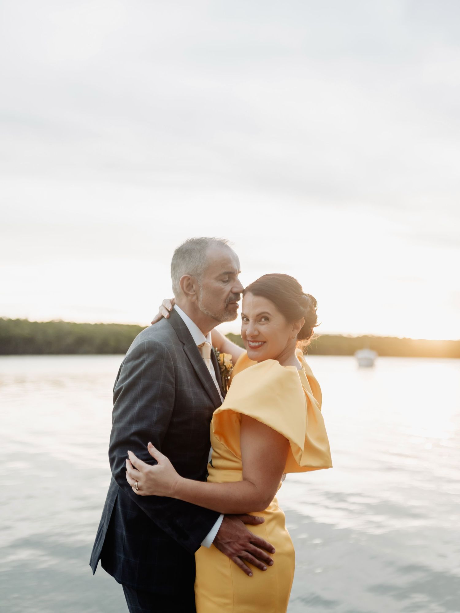 Couple embraces at golden hour by the water, bride in yellow dress and groom in navy suit sharing a kiss.
