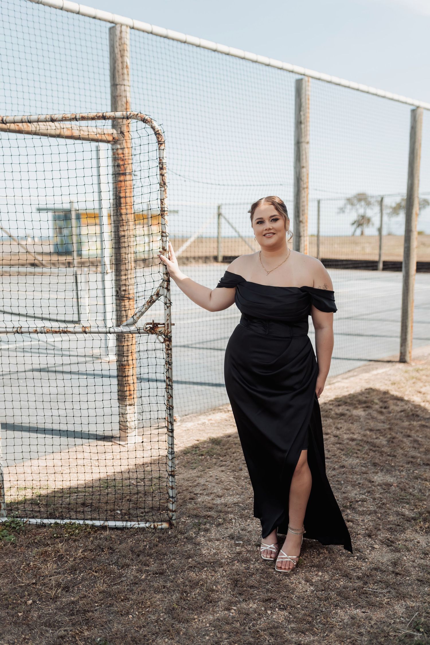 A person in a flowing black dress poses gracefully by a mesh fence near the beach.