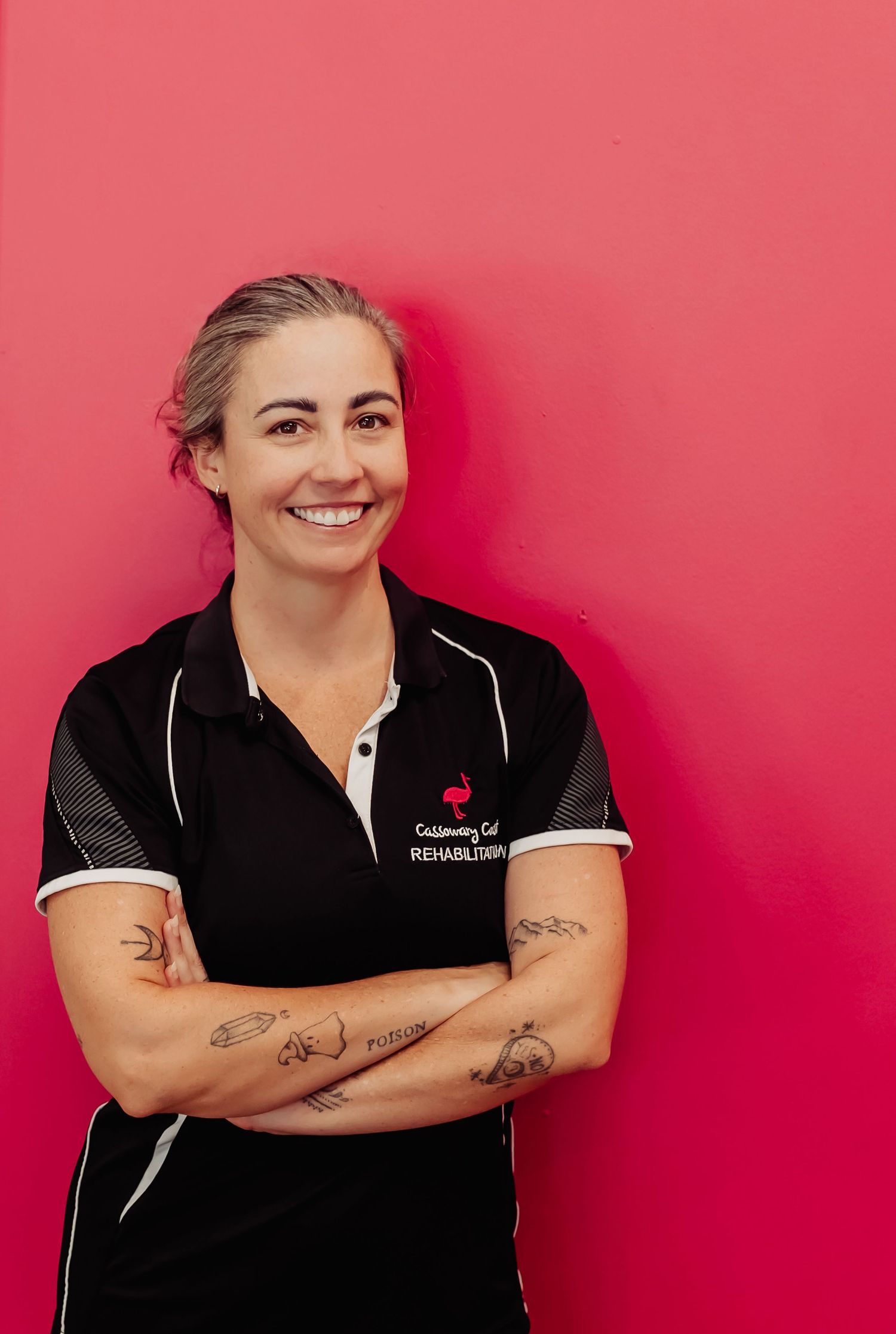 Smiling woman with tattoos in black rehabilitation polo shirt, arms crossed, standing against a bright pink wall.
