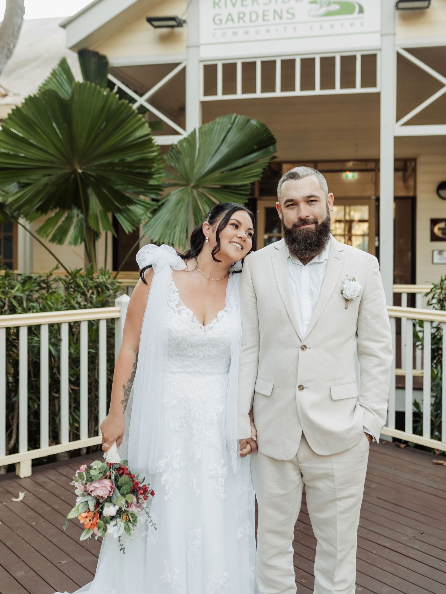Bride in white lace gown with veil and groom in cream suit pose together at Riverside Gardens venue.
