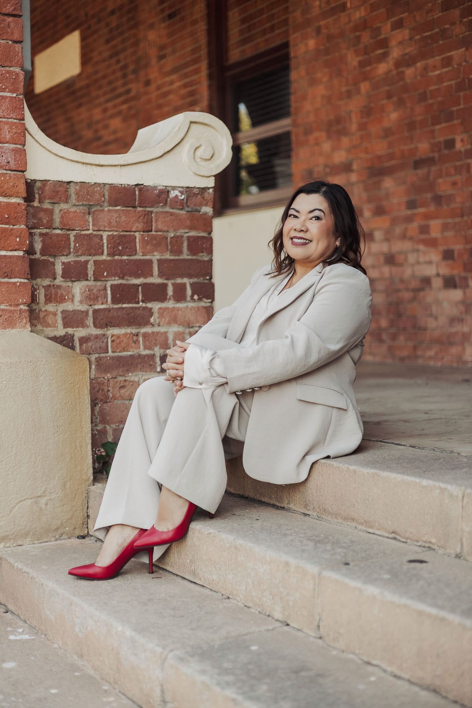 Woman in cream suit and red heels poses elegantly on brick building steps.