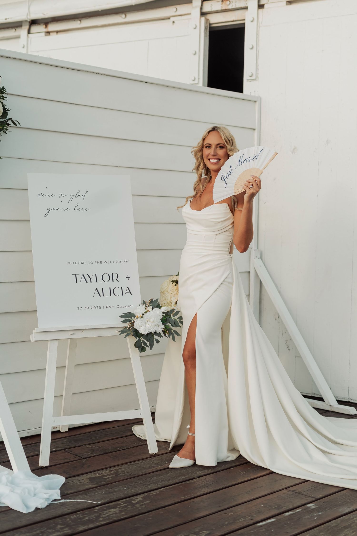 Bride poses energetically in flowing white gown against white backdrop with wedding sign.