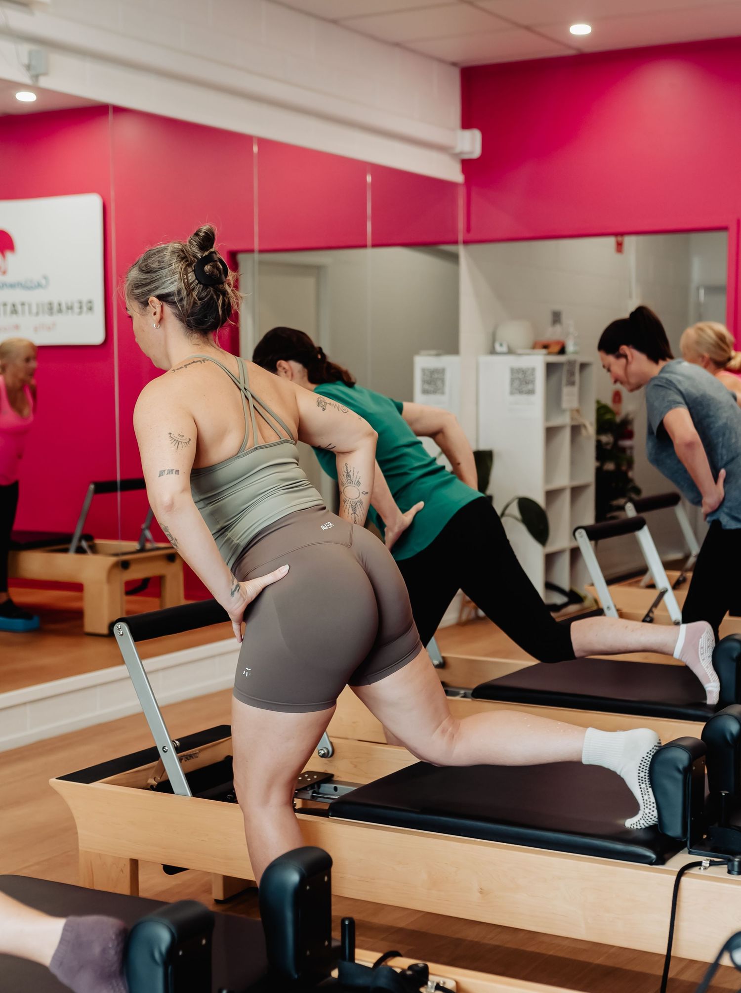 Women exercising on Pilates reformers in a bright pink studio with mirrored walls during a group fitness class.