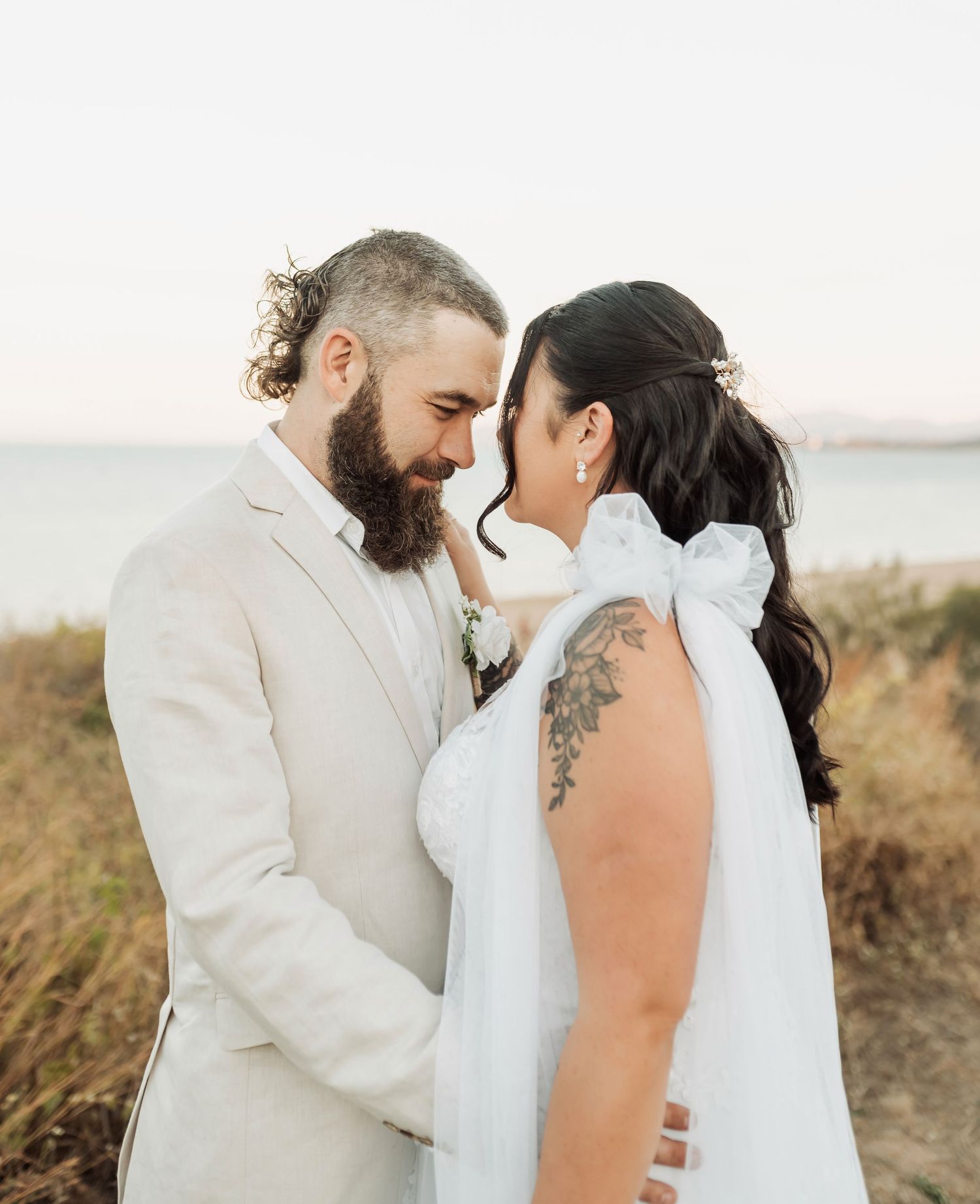 Couple in wedding attire sharing an intimate moment on a beach, groom in cream suit, bride in white gown with bow.