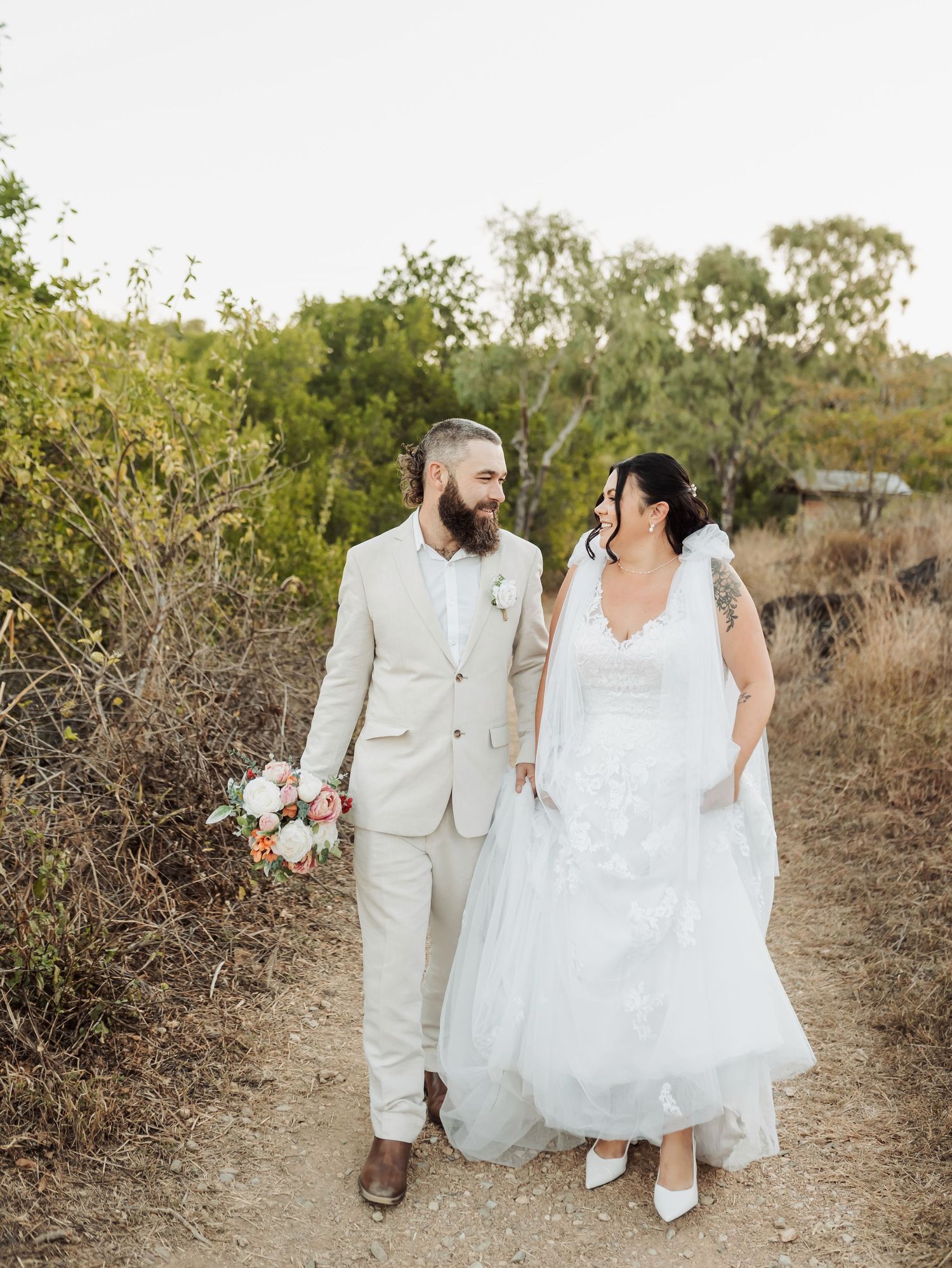 Bride and groom walk hand in hand on rustic trail, smiling at each other outdoors.