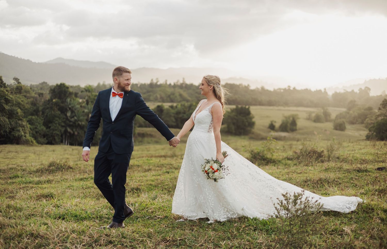 Wedding couple walks hand in hand across a scenic hilltop at sunset.