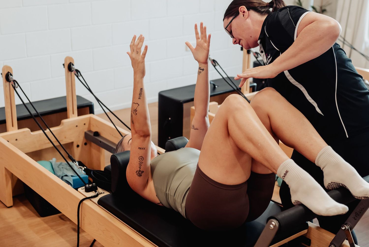 A Pilates instructor assists a client performing reformer exercises with raised arms in a bright studio.