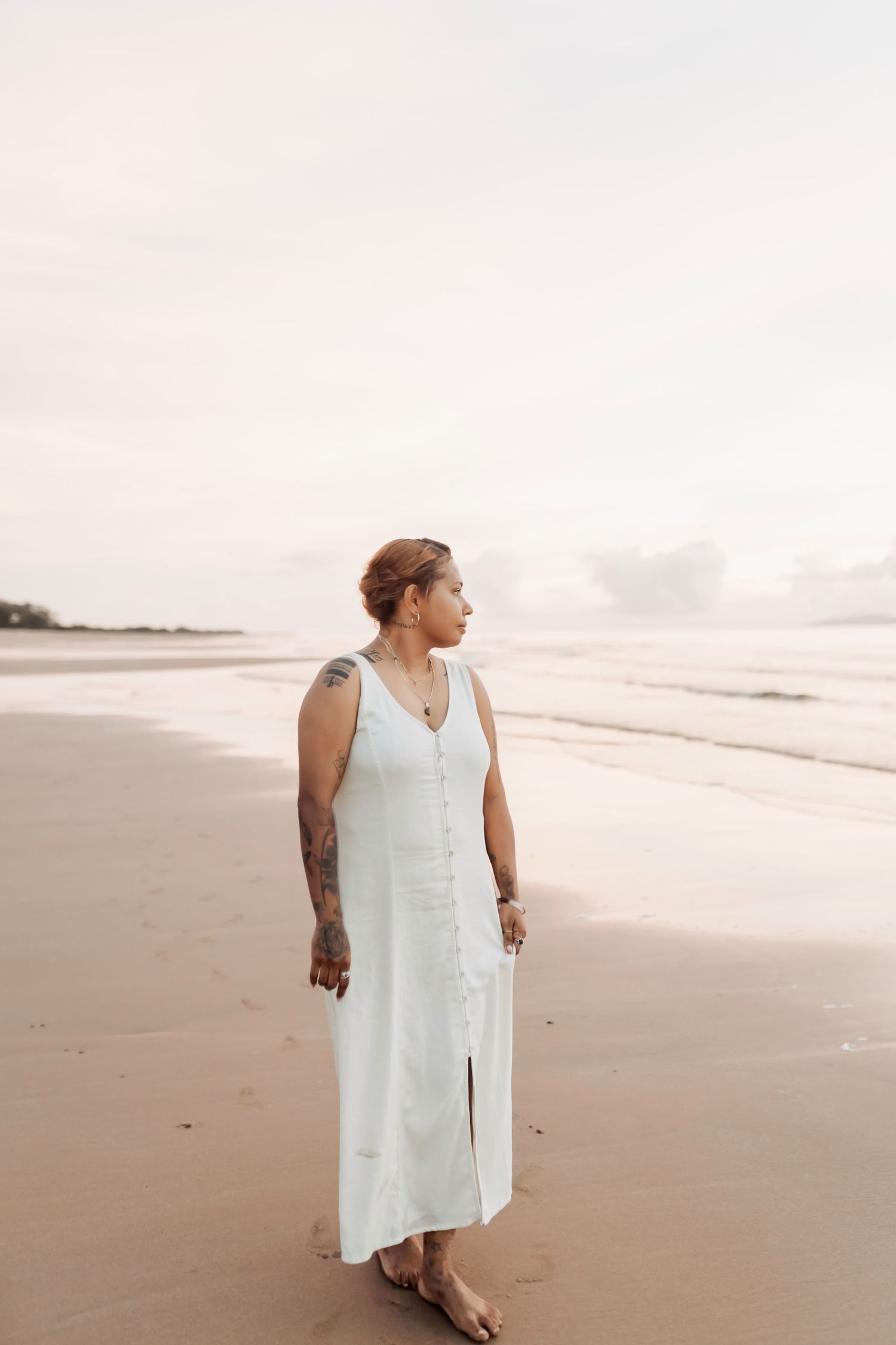 Person in white sleeveless dress poses on empty beach at dusk.