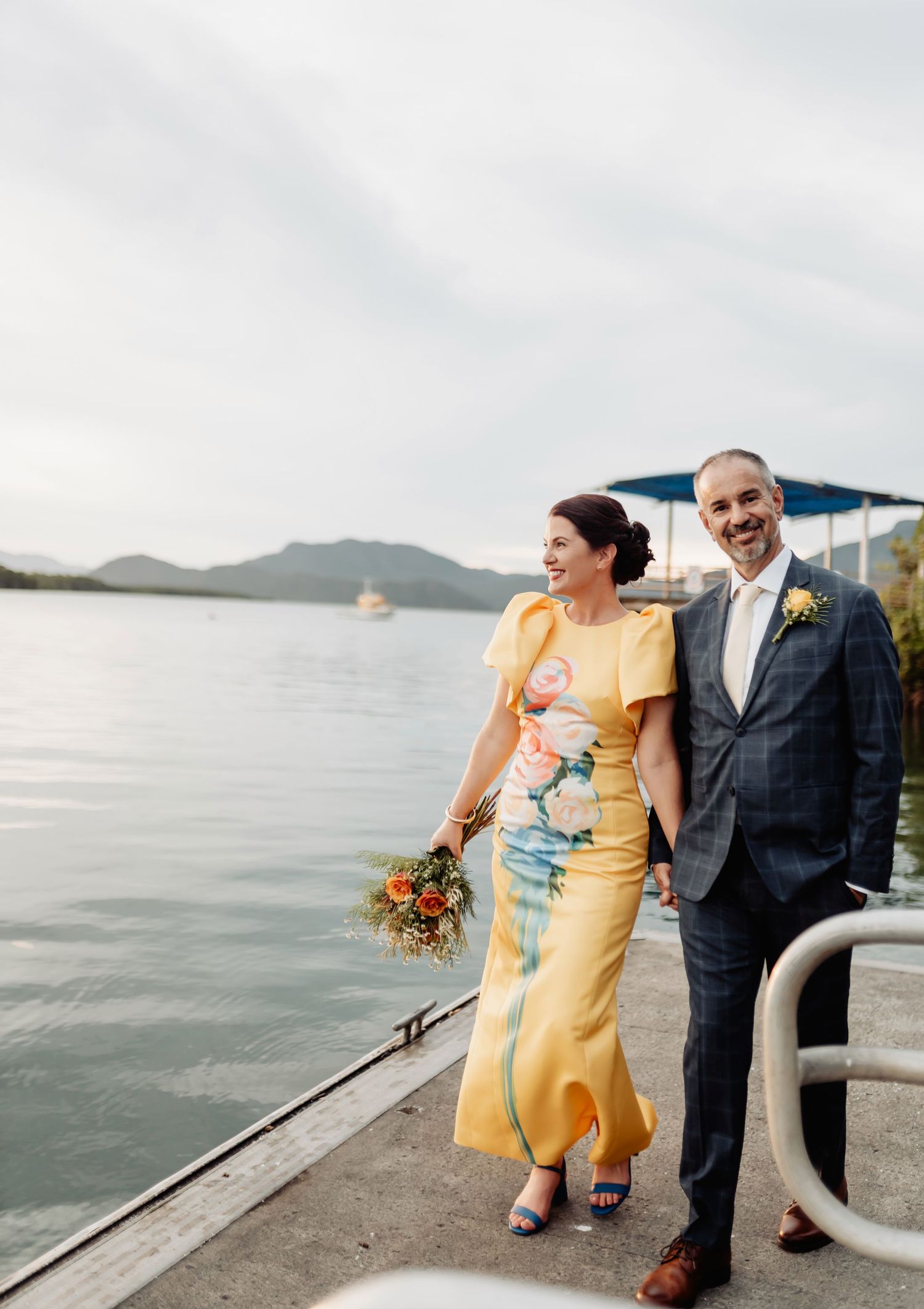 Bride in yellow floral dress holds bouquet beside groom in navy suit on waterfront dock with mountain views.