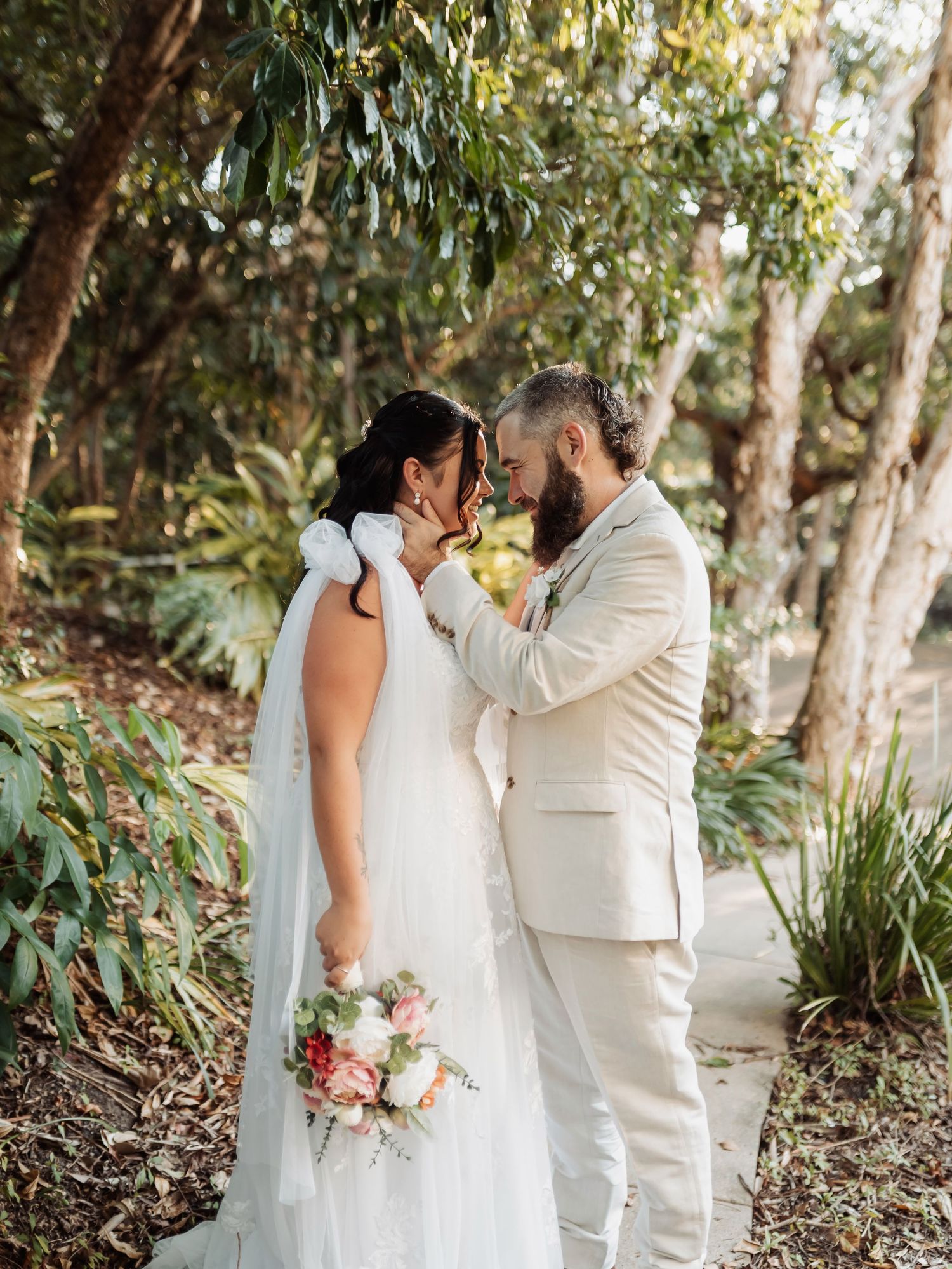 A bride and groom share a romantic moment in a lush garden, she holds a pink bouquet in a white gown, he wears a beige suit.