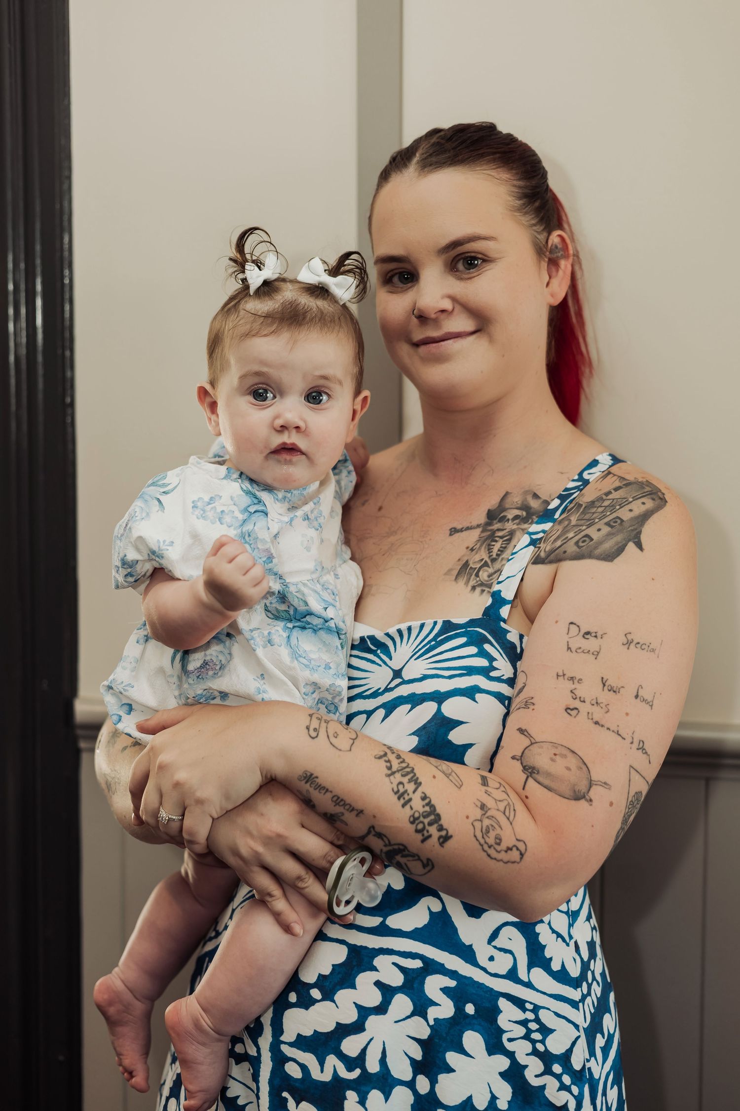 Mother in blue floral dress holding baby wearing white outfit in series of indoor portraits.