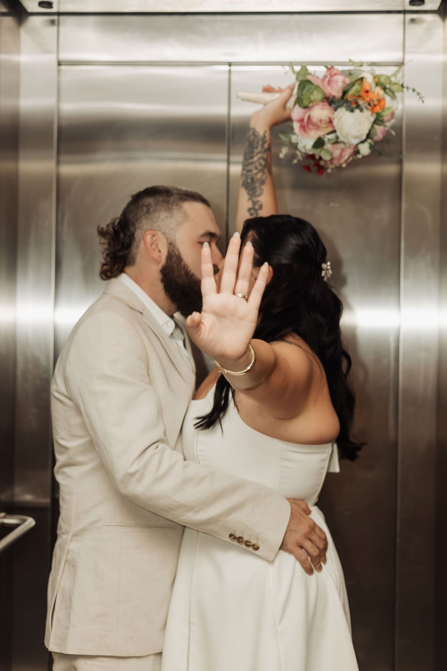 Bride and groom sharing a kiss in elevator, bride holding colorful bouquet aloft while blocking camera with her hand.