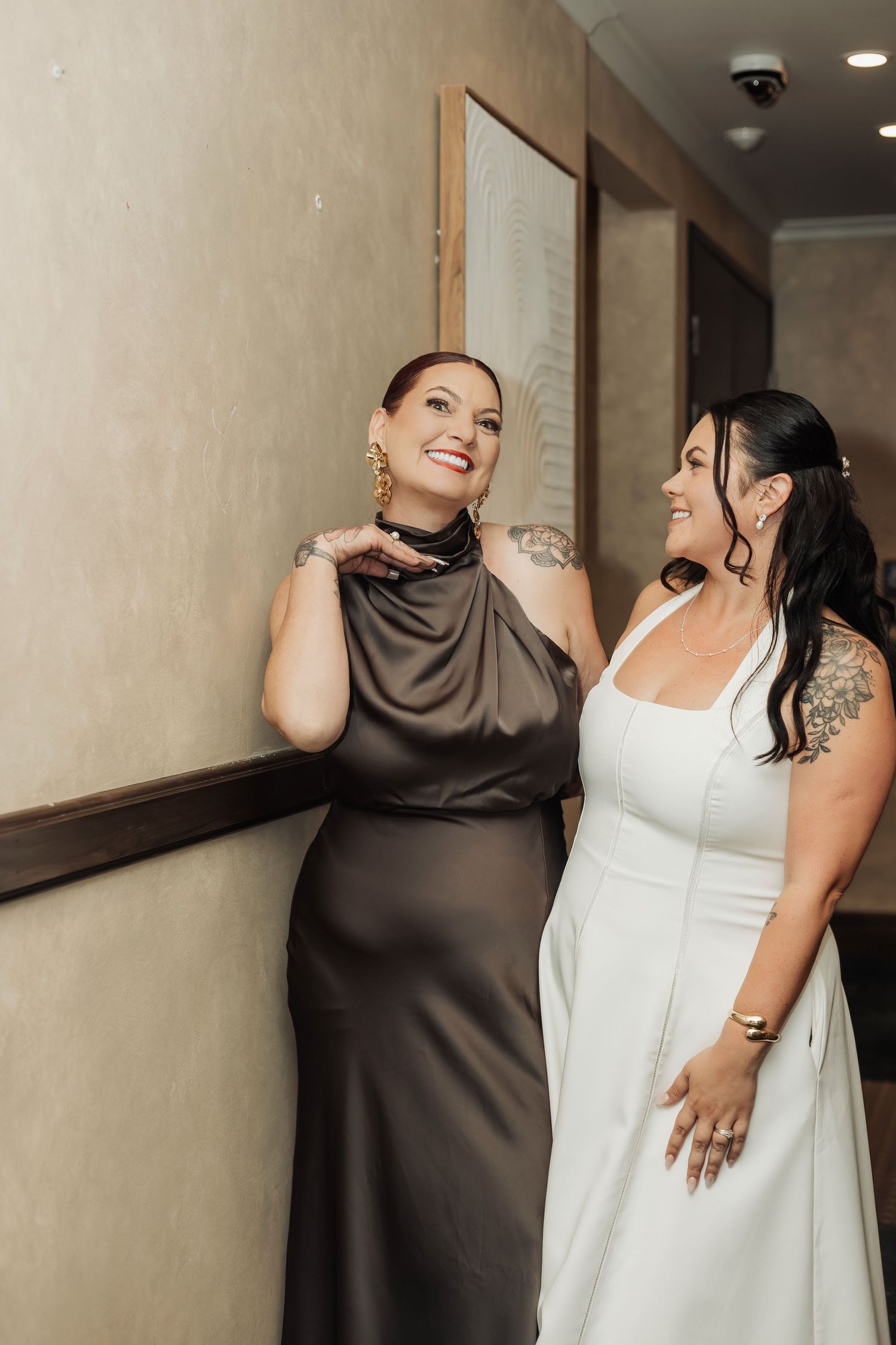 Bride in white dress and bridesmaid in brown satin gown smiling together in a hallway.
