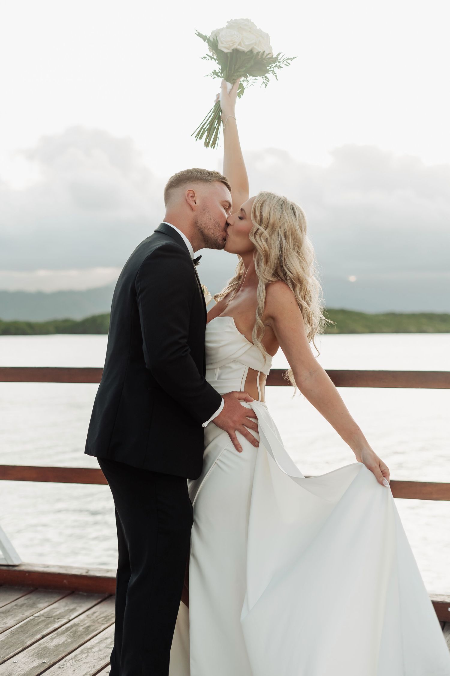 Romantic wedding couple embraces and kisses on wooden deck overlooking water.