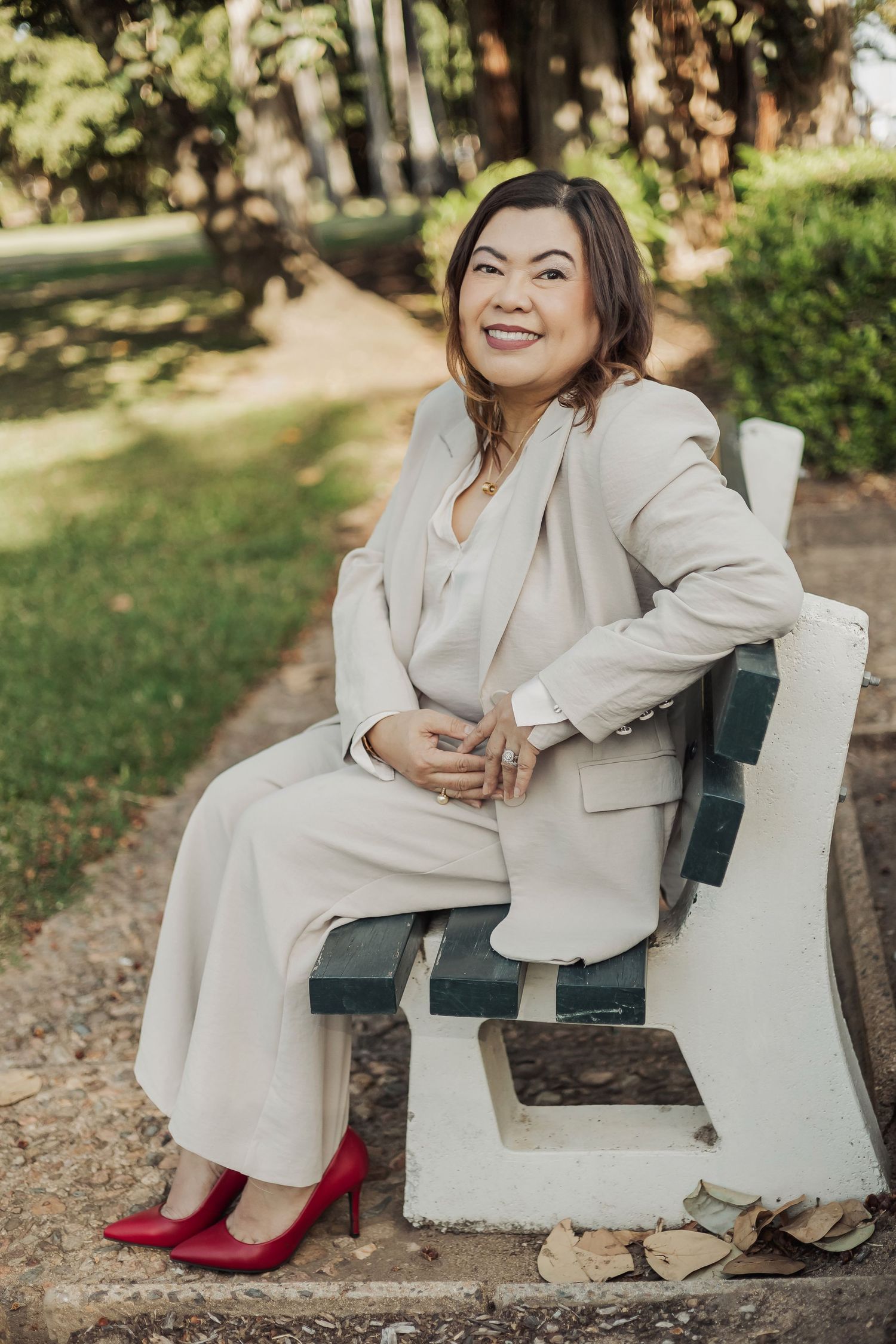 Woman in cream pantsuit sits casually on outdoor bench wearing bright red shoes.