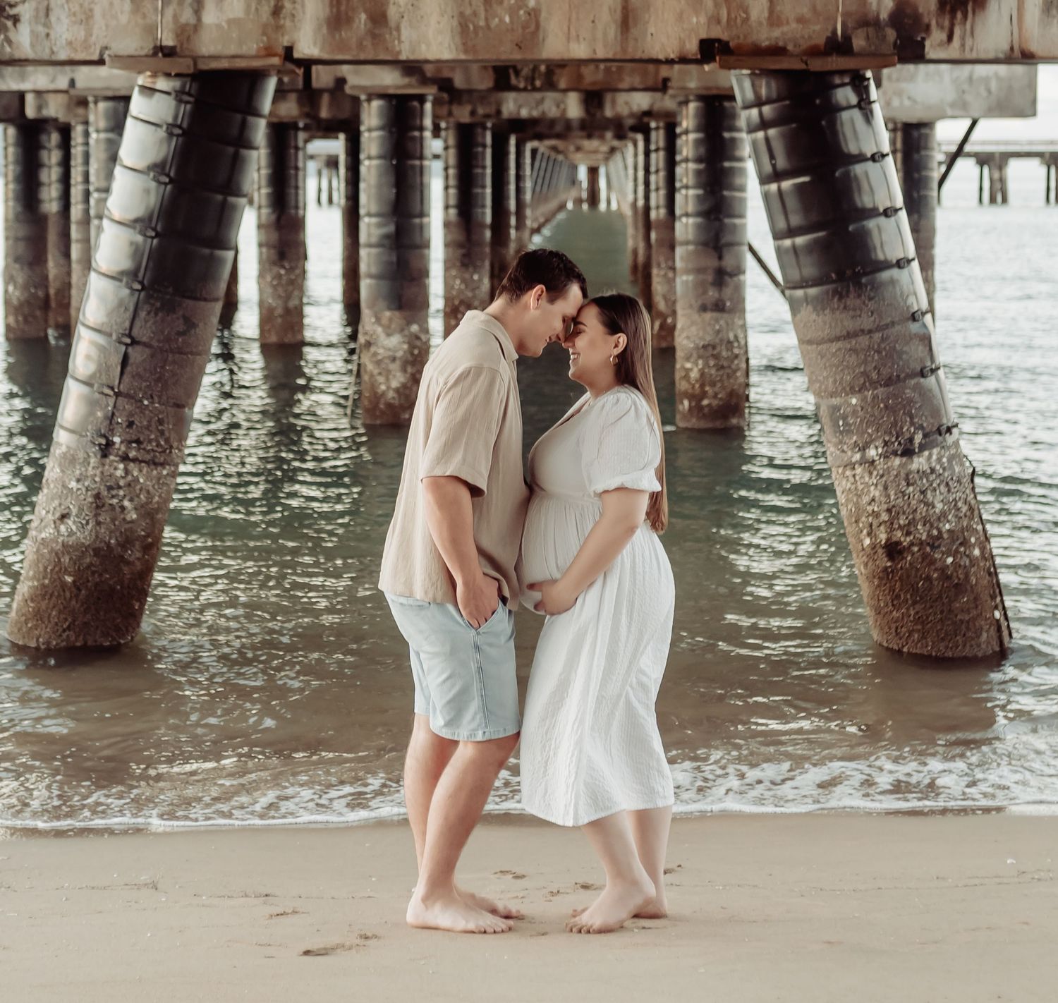 Couple sharing a tender moment on sandy beach under a pier, woman in white maternity dress, man in beige shirt and blue shorts.