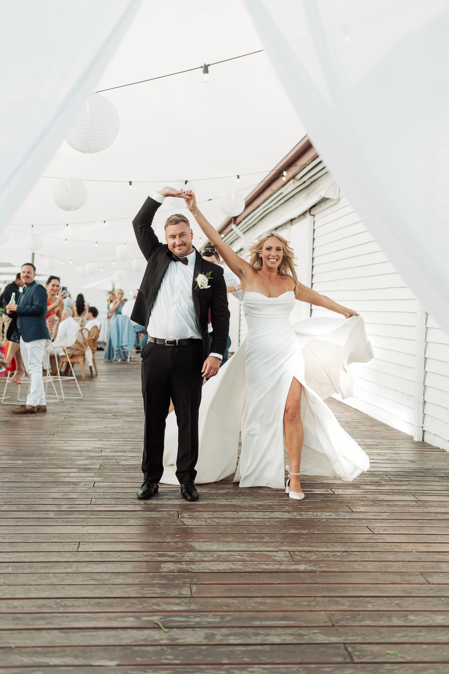 Playful wedding couple dances on wooden boardwalk under white tent.