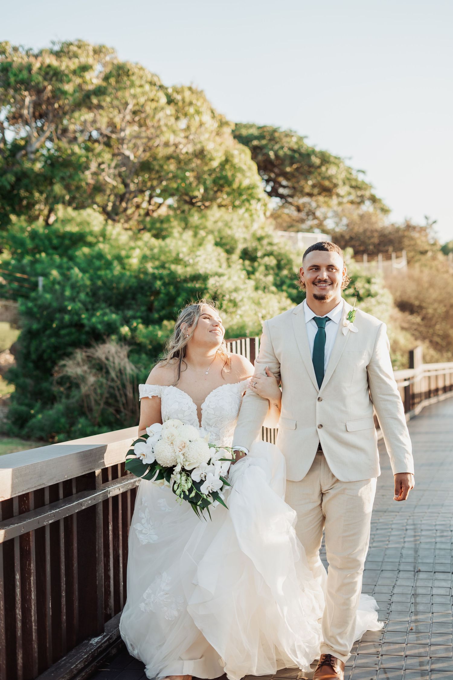 A couple in wedding attire shares joyful moments walking along a wooden boardwalk surrounded by lush greenery.