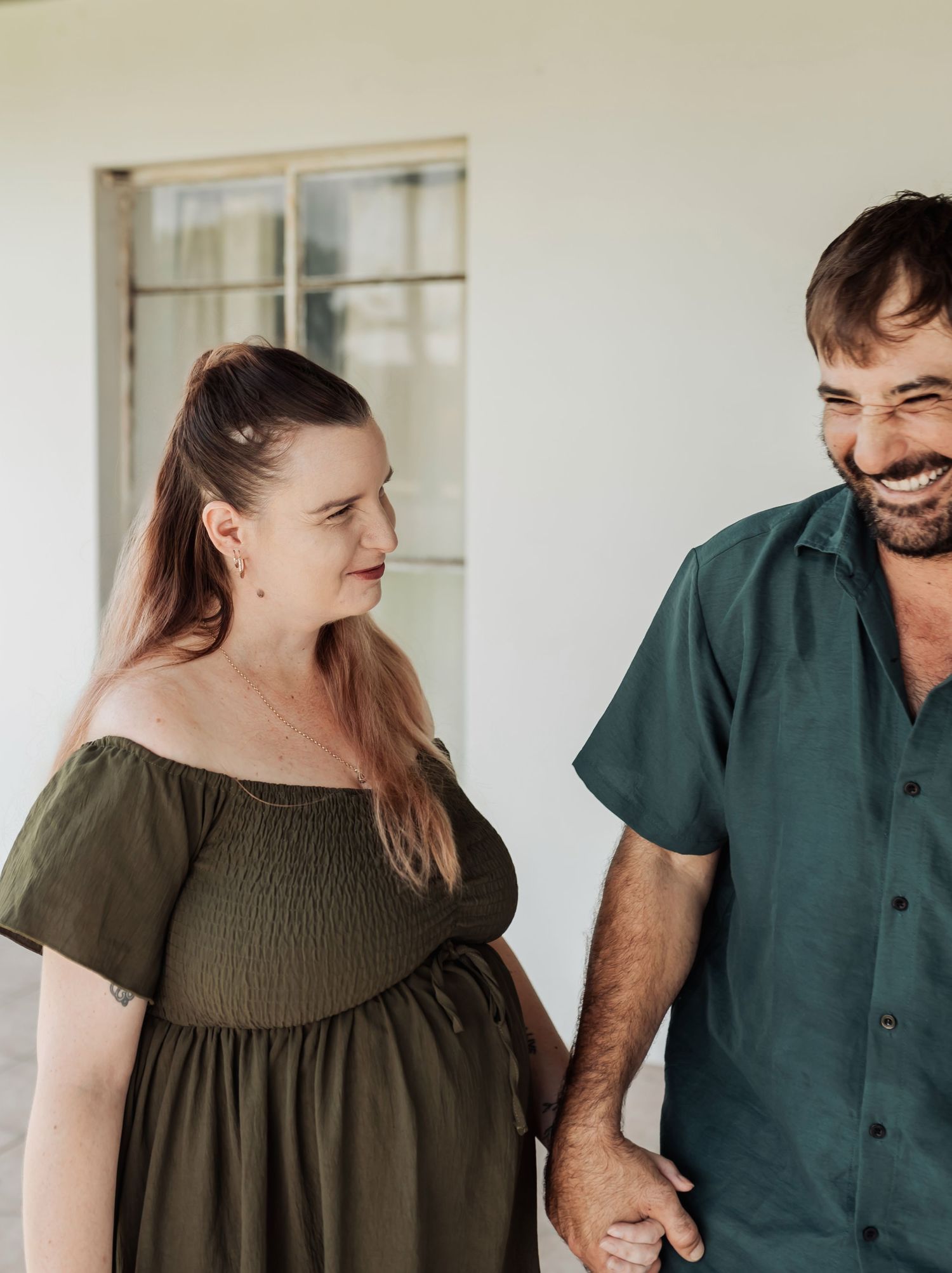 Smiling couple holding hands, pregnant woman in olive off-shoulder dress with partner in teal shirt.