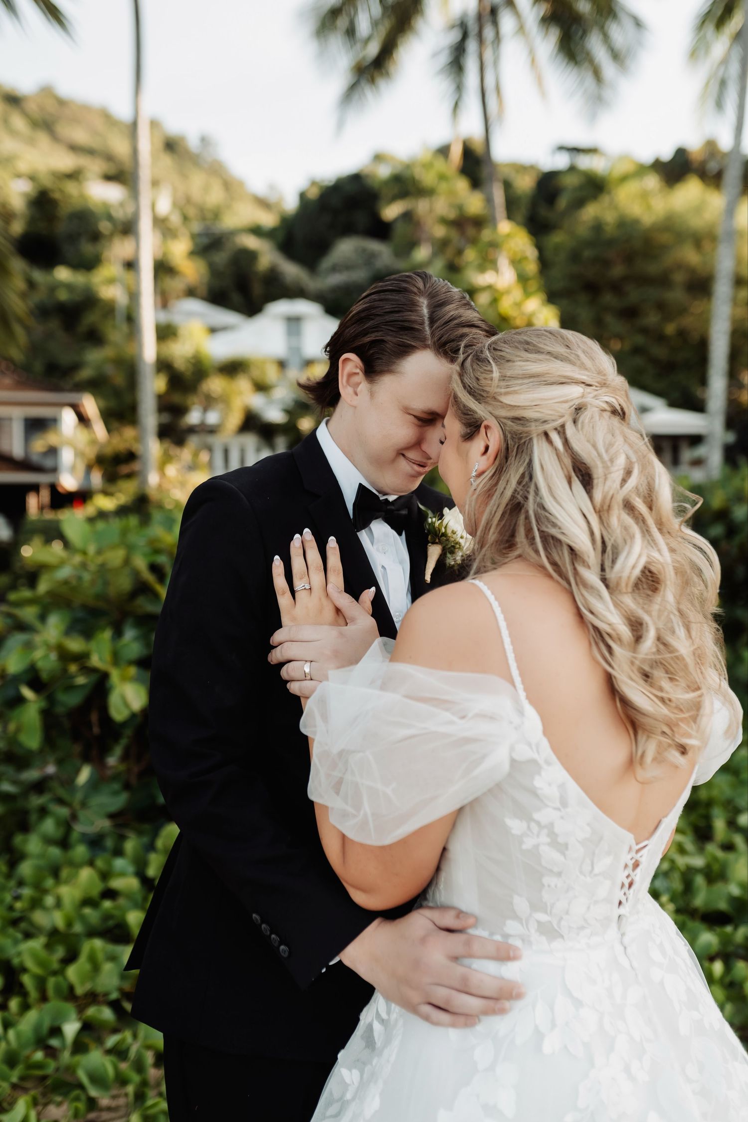 Bride and groom in wedding attire share an intimate forehead touch outdoors with tropical greenery background.