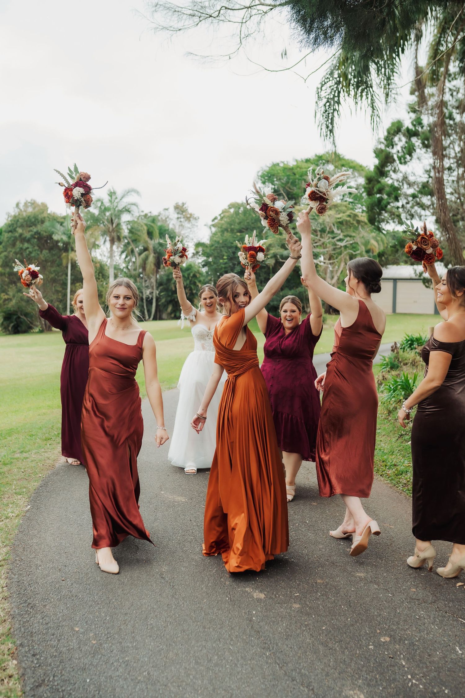 Wedding party in burgundy and rust-colored dresses celebrating on a garden path.