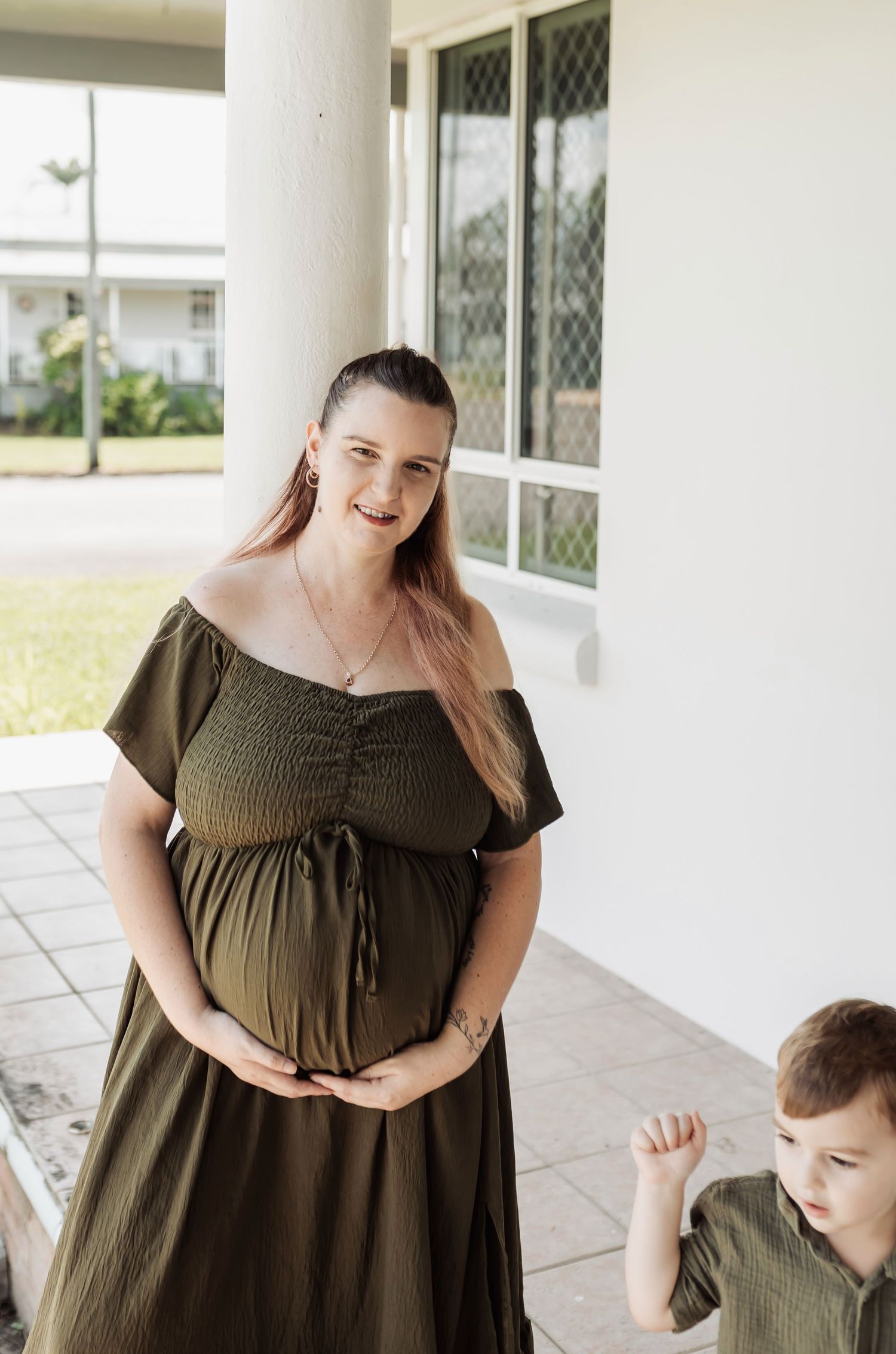 Smiling pregnant woman in olive green off-shoulder dress cradling baby bump, standing on porch with young child beside her.