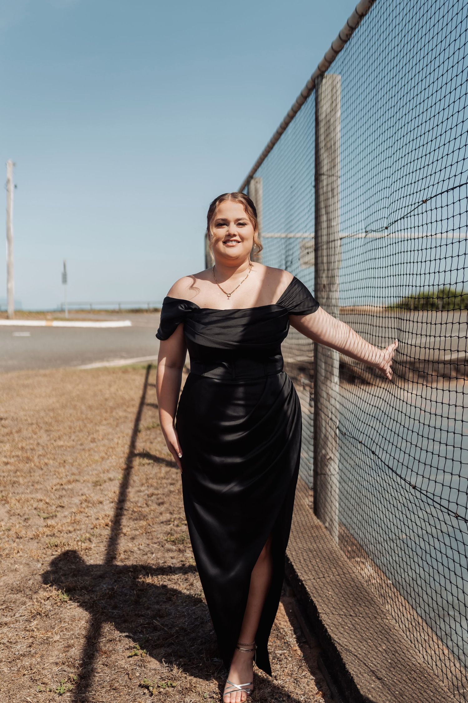 A person in a black dress poses confidently next to a mesh fence in bright sunlight.