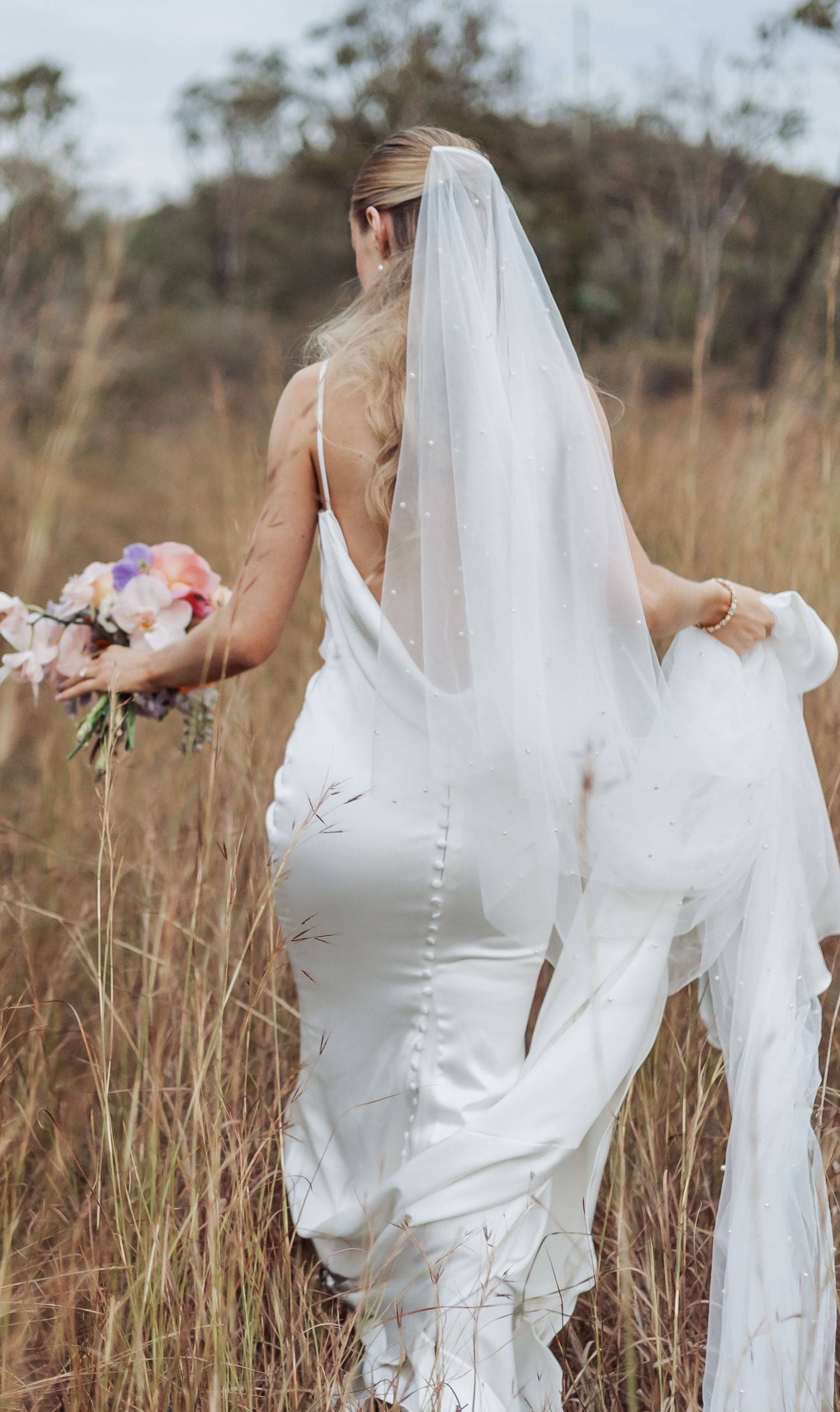 Bride in flowing white dress walks through tall grass field with flowing veil.