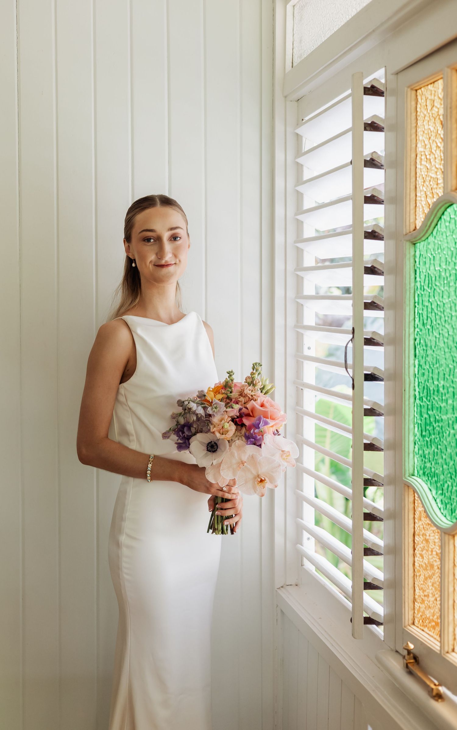 Bride holding vibrant bouquet poses by window with colored glass panels.