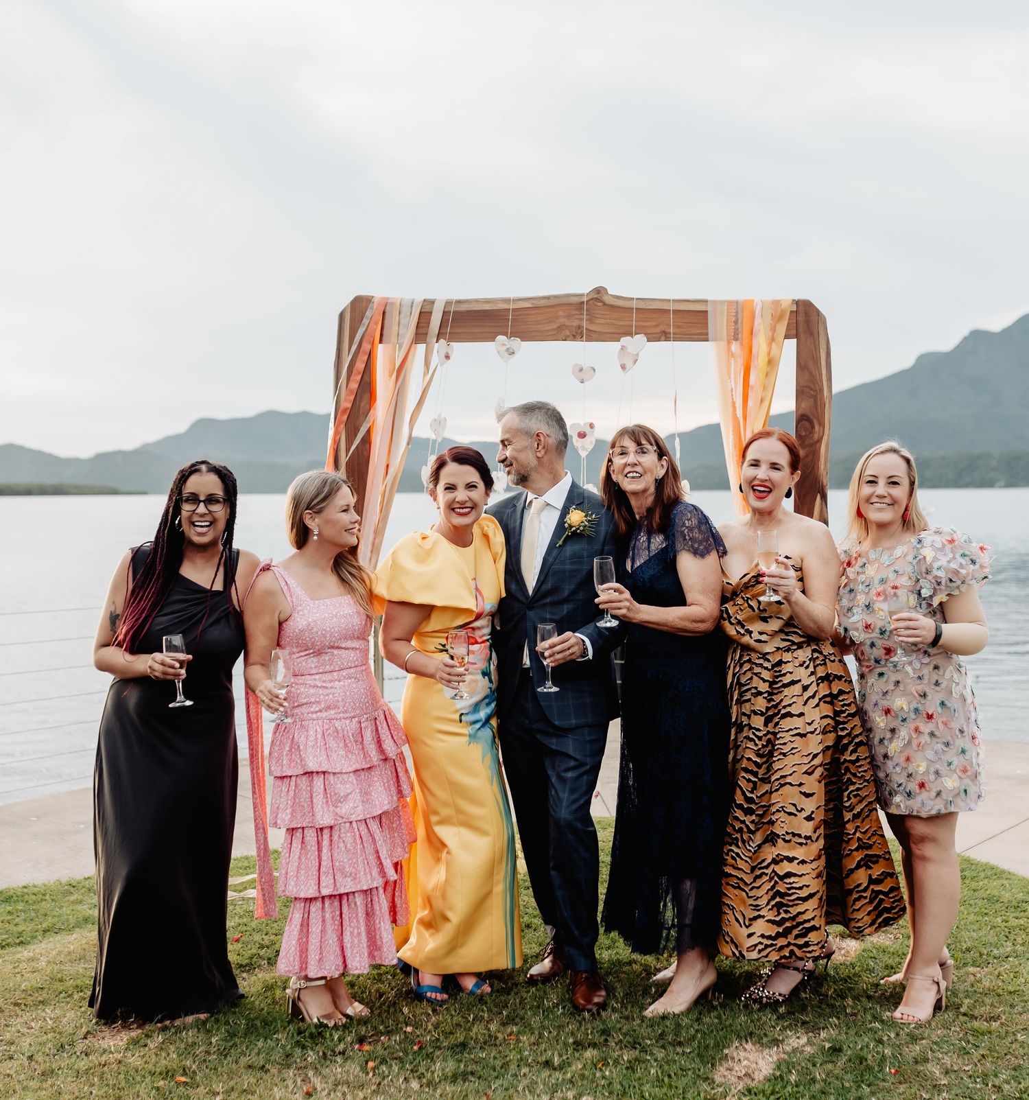 A joyful wedding party poses outdoors by a lakeside under a wooden arch with colorful ribbons.