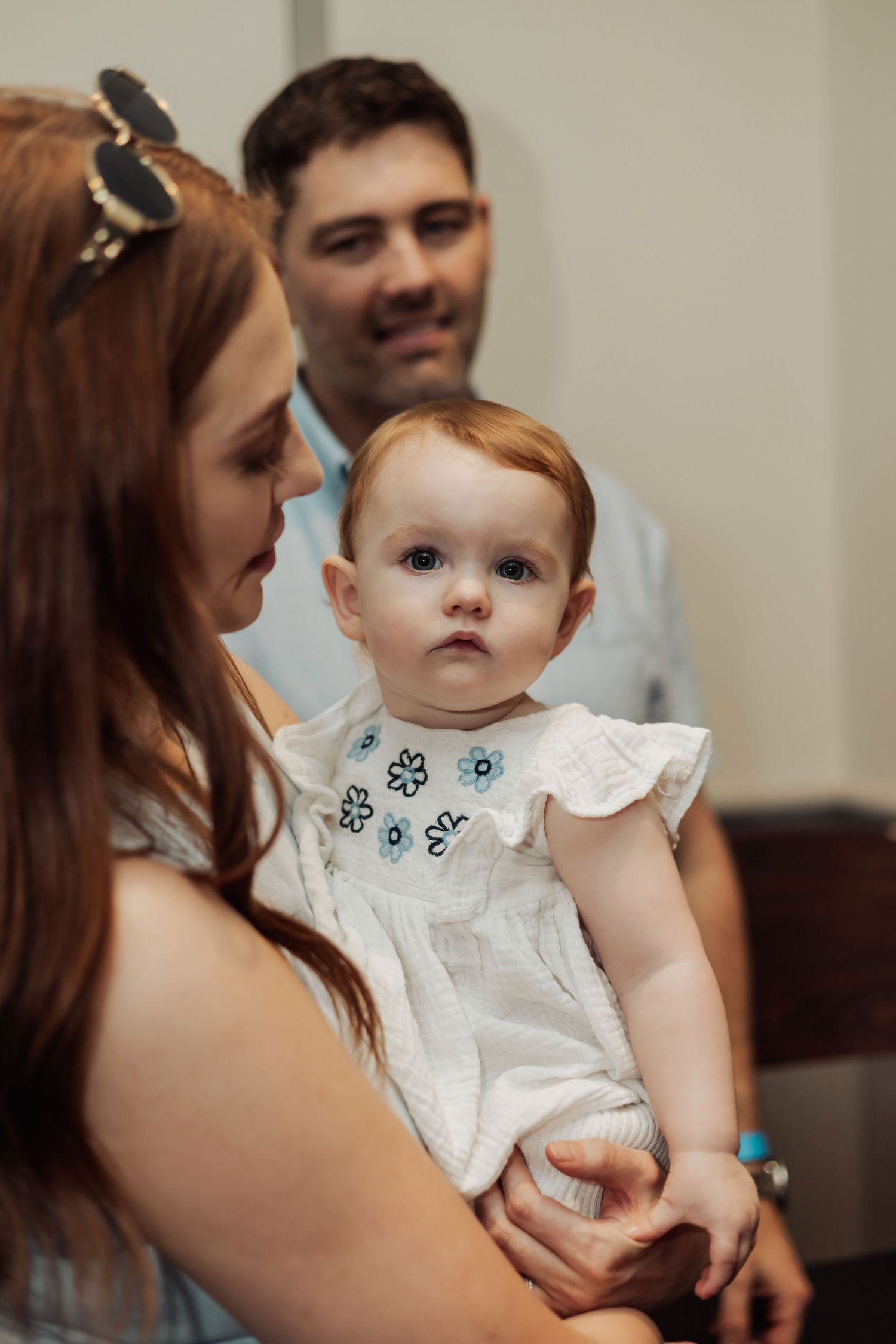 Baby in white dress being held and admired in indoor setting.