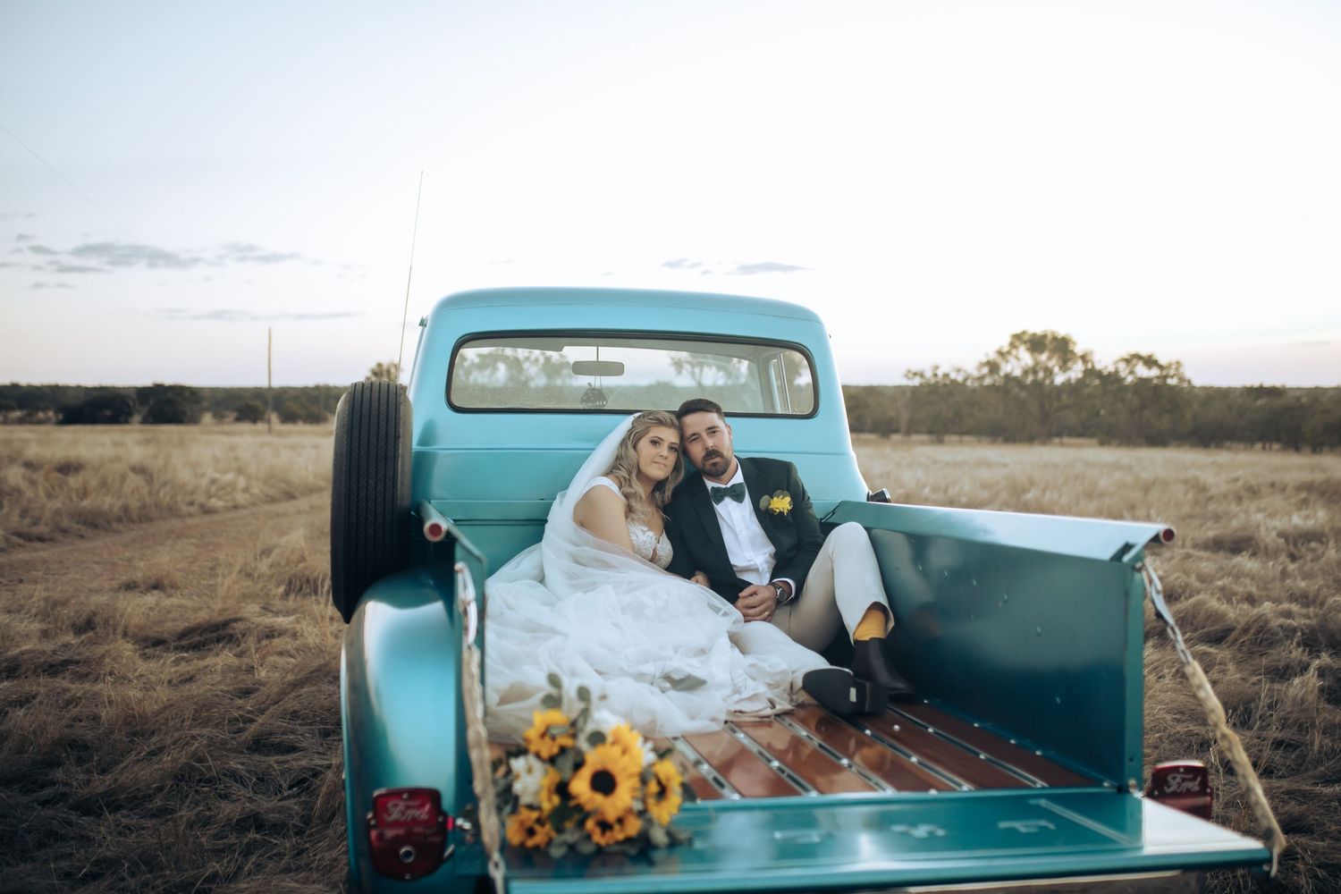 Newlyweds pose with a vintage turquoise pickup truck surrounded by sunflowers in a field. Long Horns Charters Towers