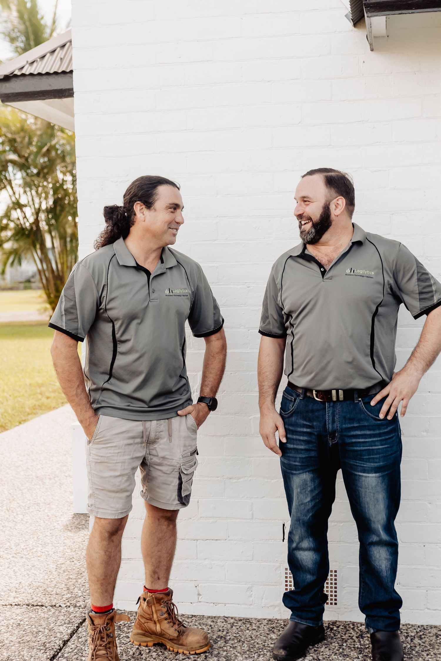 Two men in grey polo shirts laughing together outside a white brick wall.