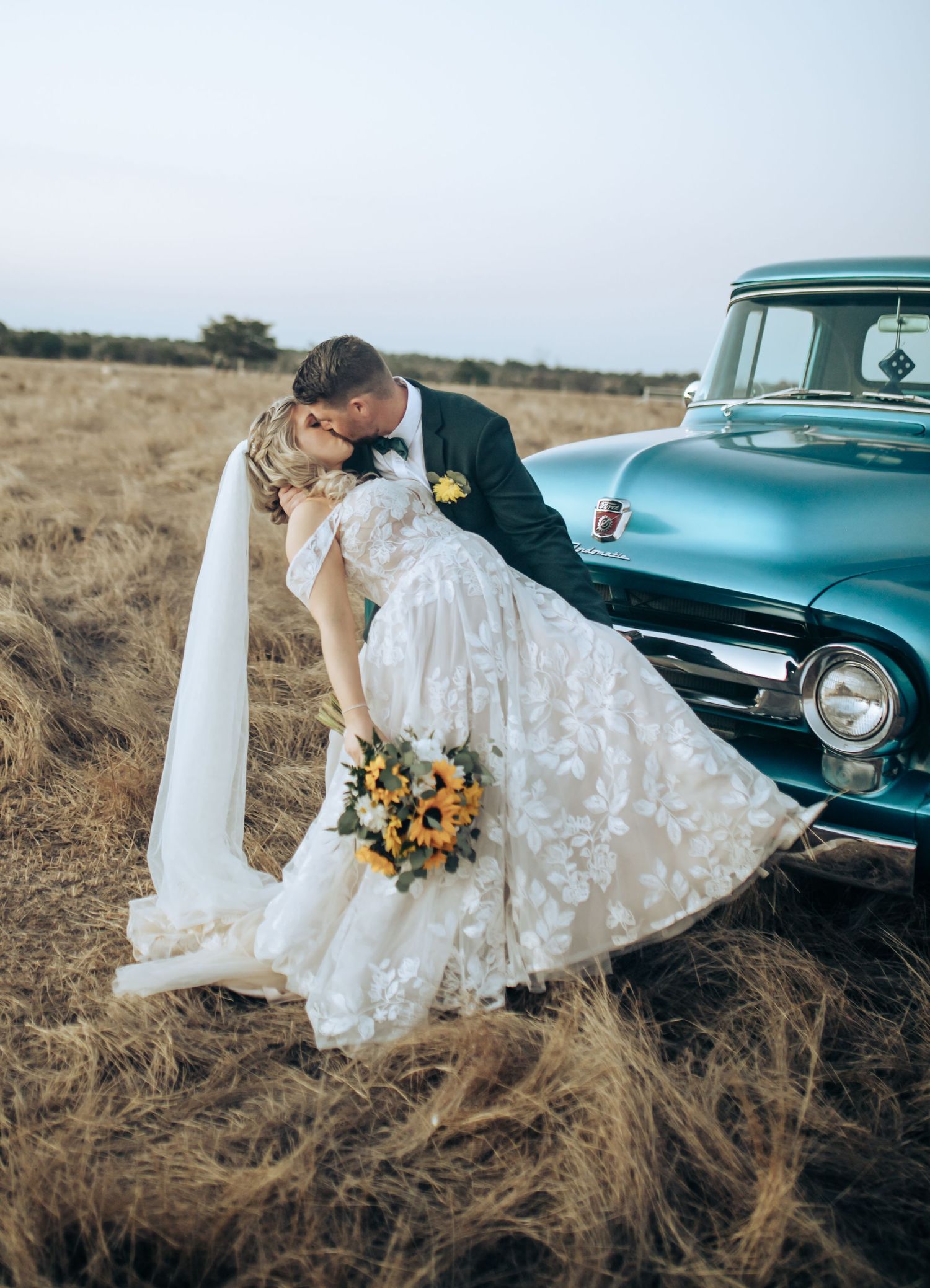 Bride and groom embrace near a classic turquoise car in a rustic field setting. Long Horns Charters Towers