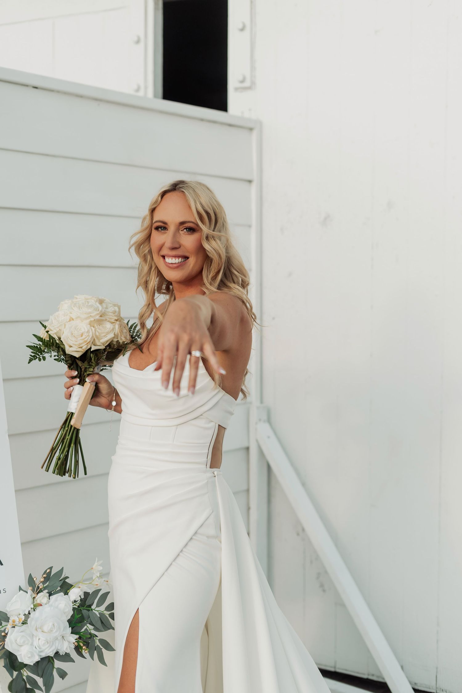 Modern bride in fitted white gown shows off bouquet against white building backdrop.