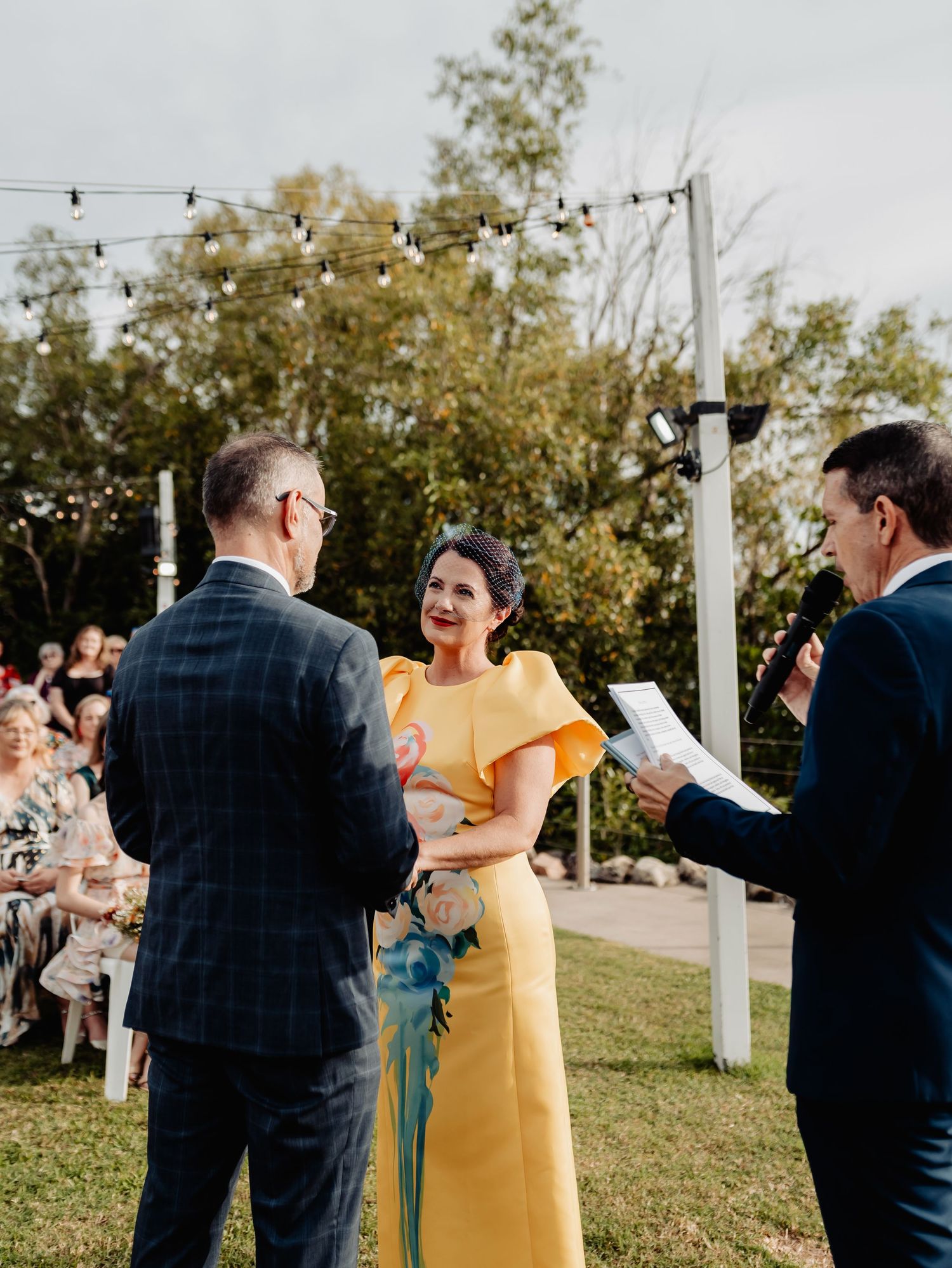 Bride in yellow dress laughing with groom and officiant during outdoor wedding ceremony with string lights.