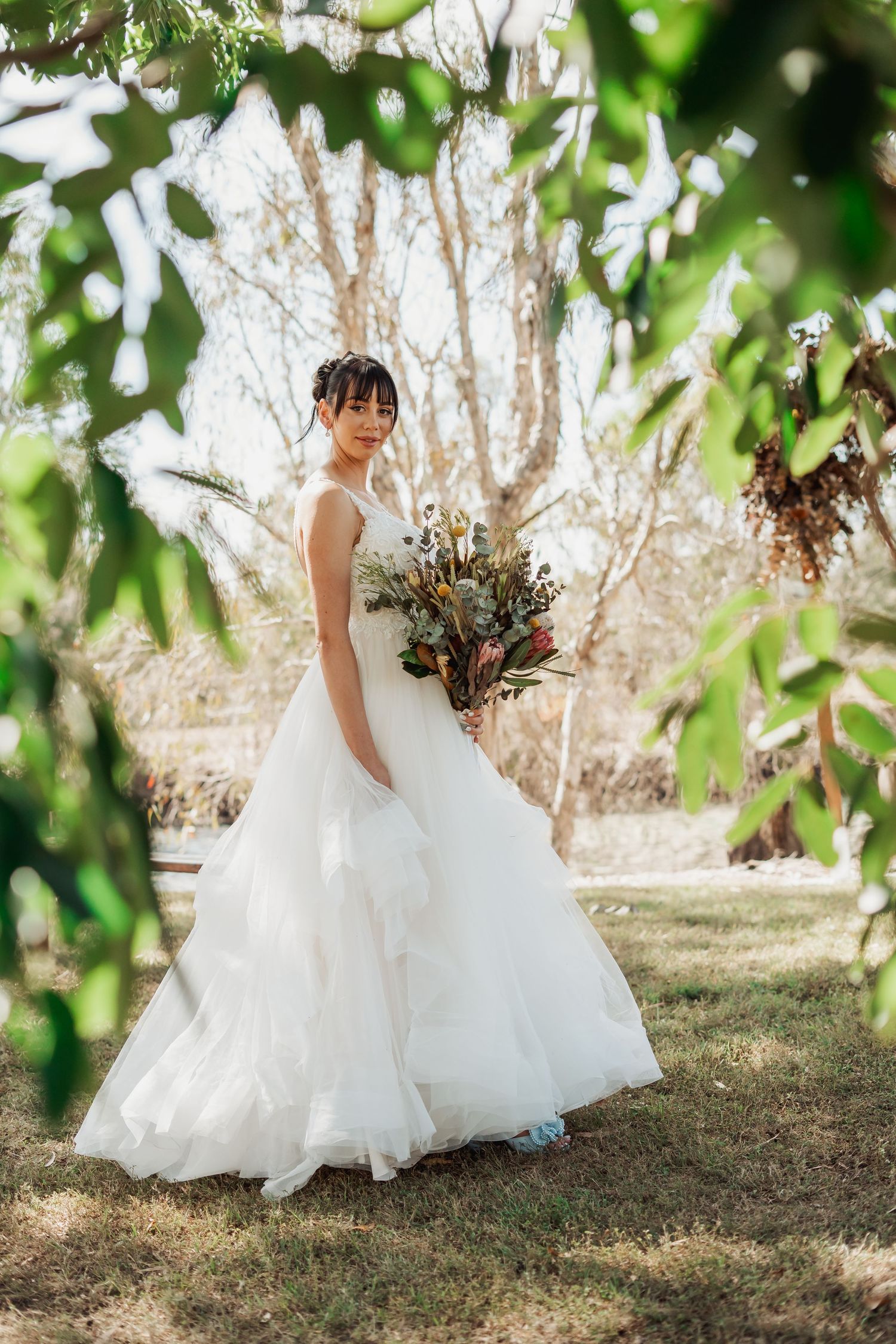 A bride in a flowing white wedding dress twirls gracefully in a sunlit garden setting with green foliage in the background. Townsville