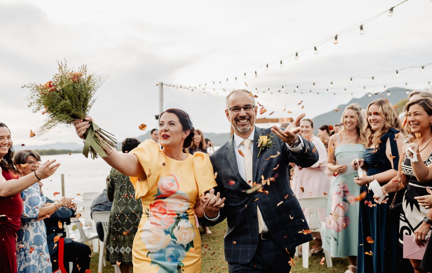 Joyful couple exits wedding ceremony as guests throw colorful confetti outdoors near waterfront with string lights.