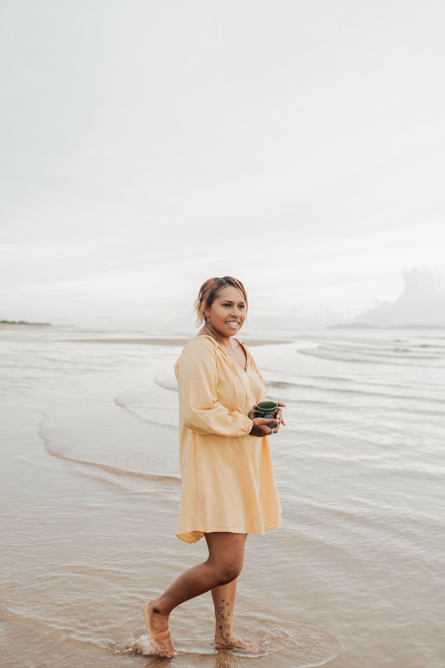 Person wading in ocean waves wearing oversized yellow shirt while holding camera.