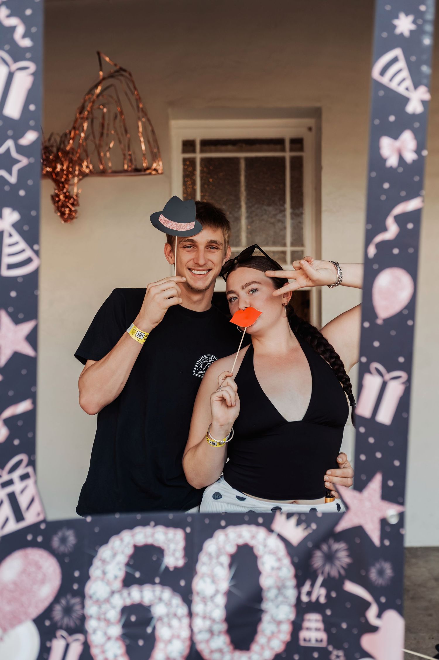 A couple poses with party props in front of a 60th birthday decorative frame.